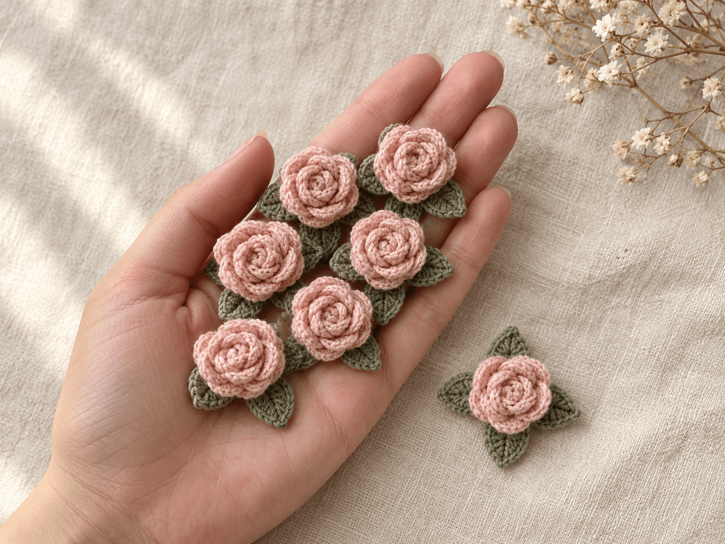 Seven blush pink crochet mini roses with sage green three-leaf bases resting on an open palm on cream linen