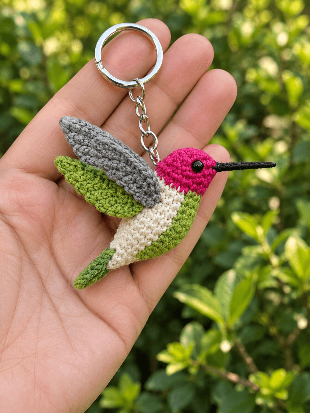 A handmade mini crochet hummingbird keychain held in the palm of a hand — a bright magenta and cream amigurumi hummingbird about 7 cm long with a green under-wing and belly, a grey upper wing, a small green tail feather and a long straight black beak, mounted on a silver split-ring keychain, soft blurred green foliage background in warm afternoon light