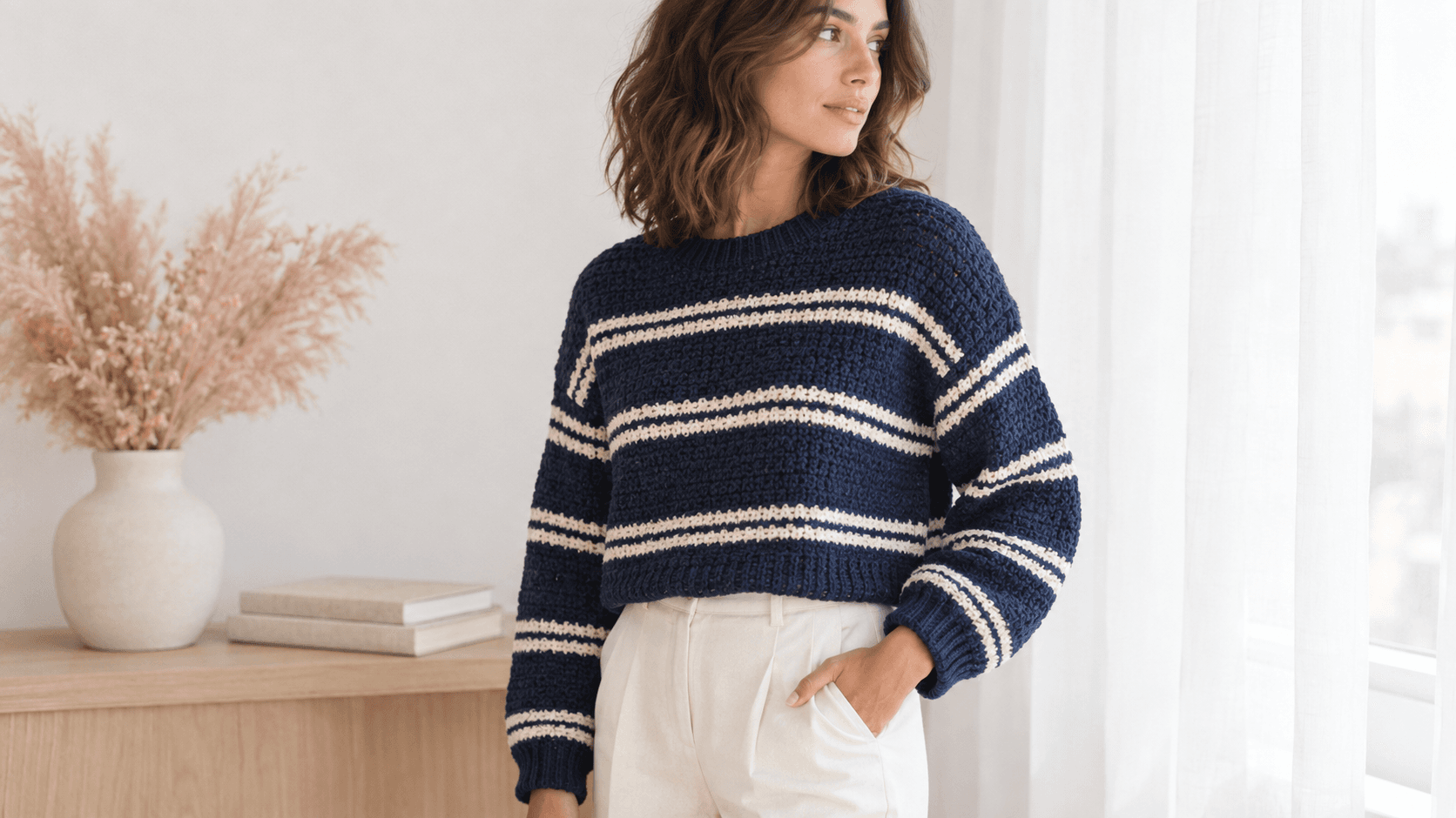 Cropped oversized crochet pullover sweater in navy blue and cream horizontal stripes, worked in half double crochet, worn by a young woman in cream trousers with dropped shoulders and ribbed cuffs, styled indoors near a bright window